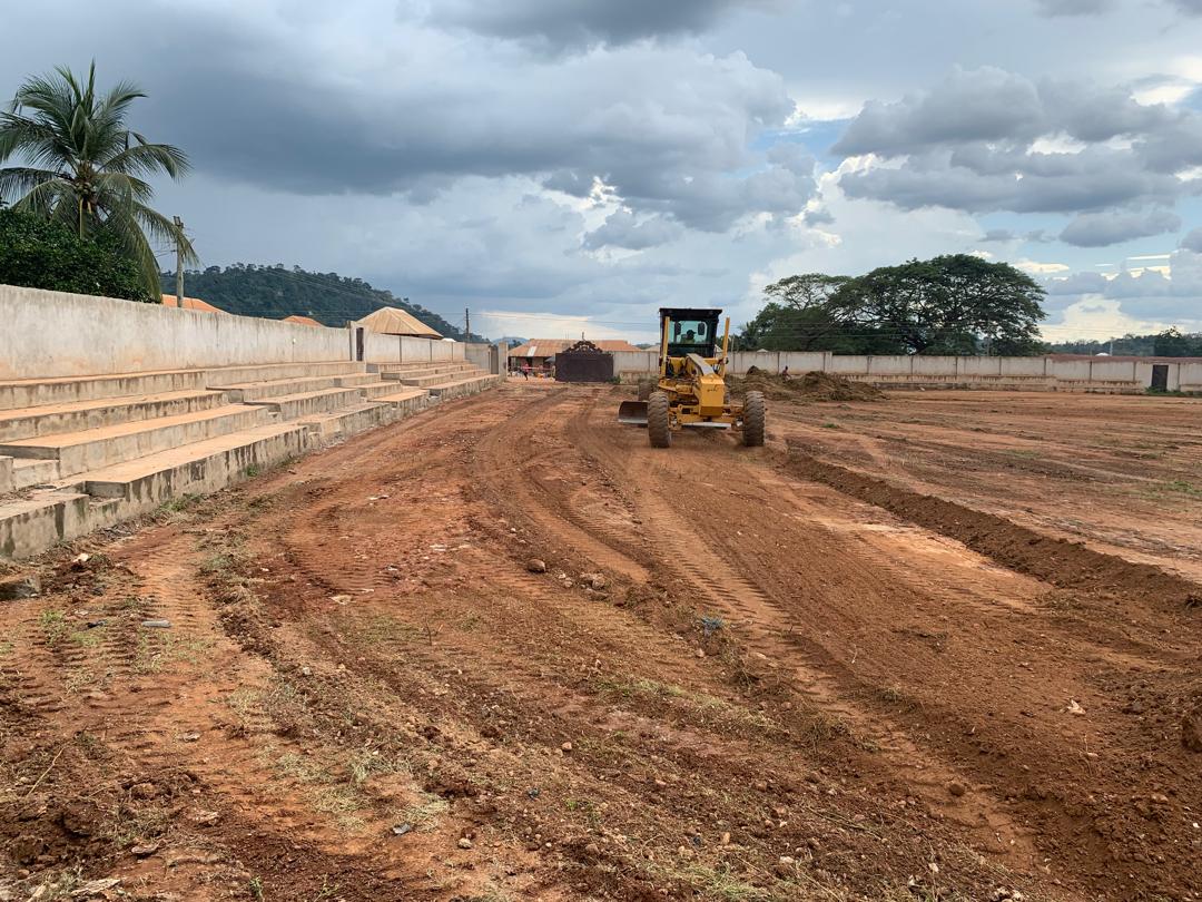 Motor grader leveling earth at a civil works site with tiered concrete works and hills in the background